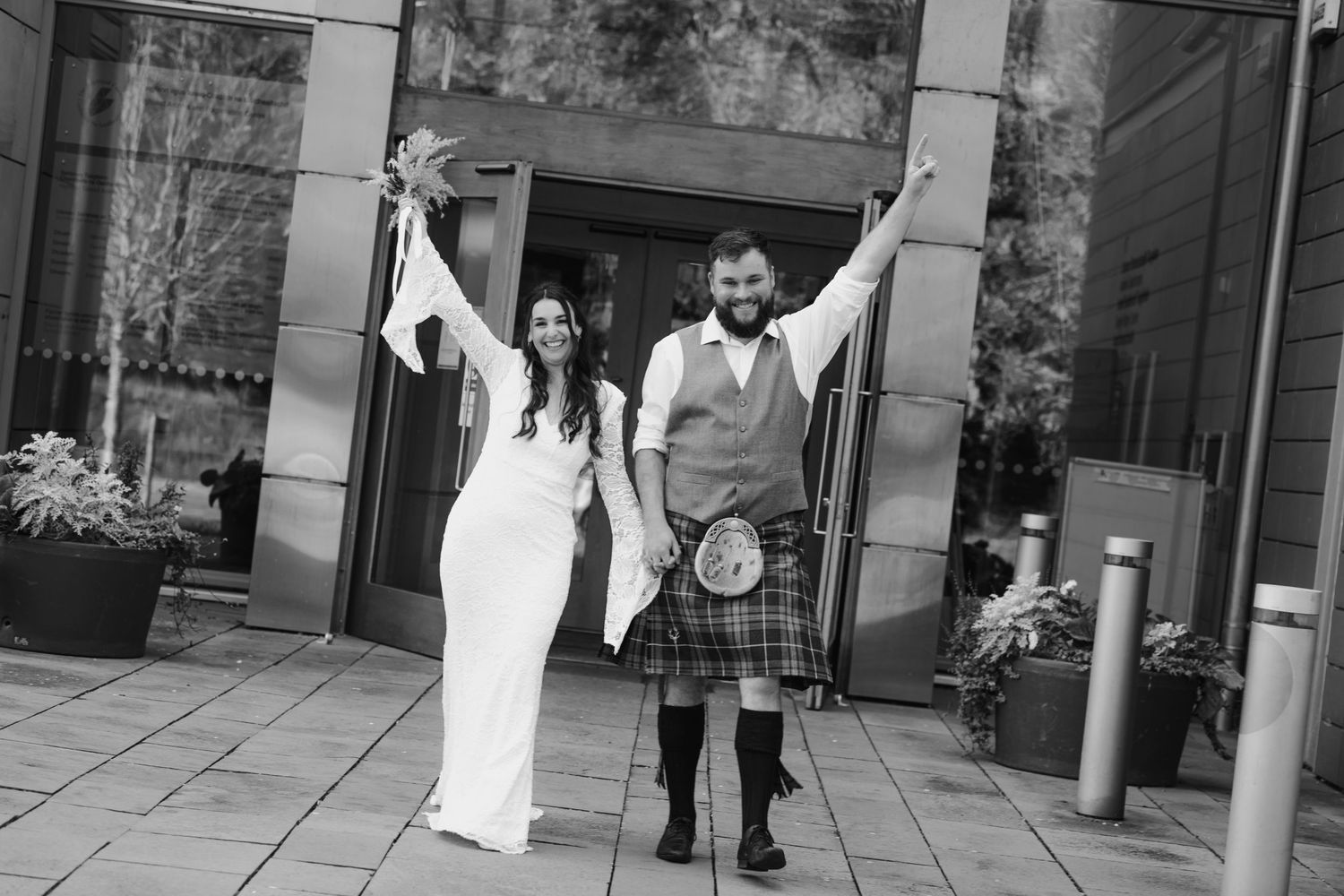 Jubilant newlyweds raise arms in celebration outside modern building in black and white portrait.