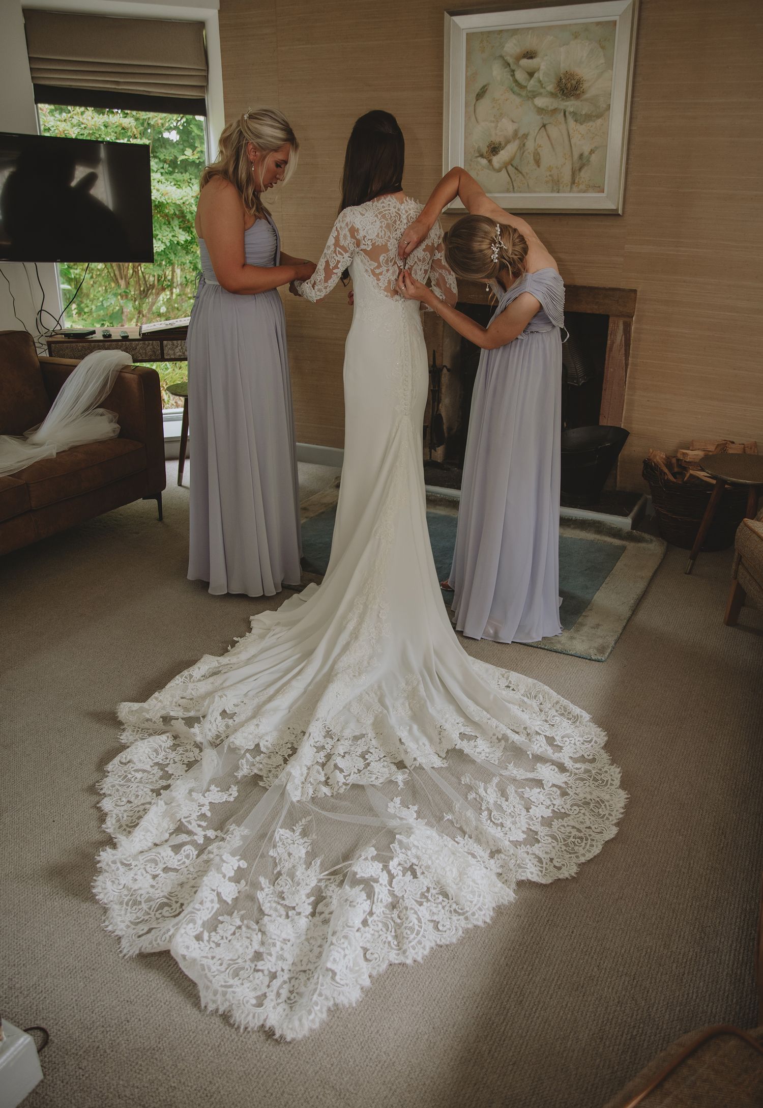 Bride in lace wedding gown with train being helped by two bridesmaids in grey dresses during wedding preparations.