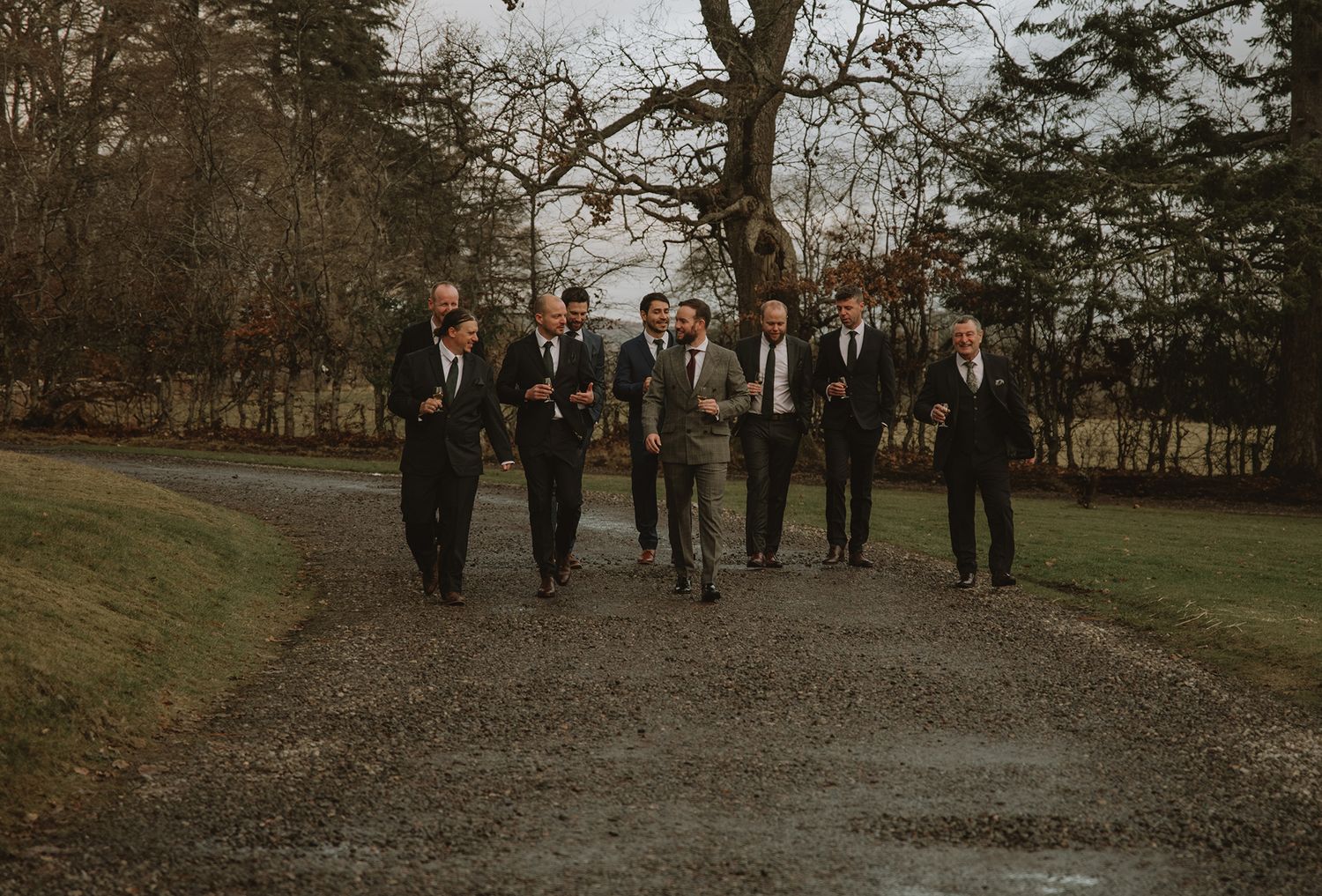 Group of men in suits walking together on a gravel path surrounded by trees and green grass.