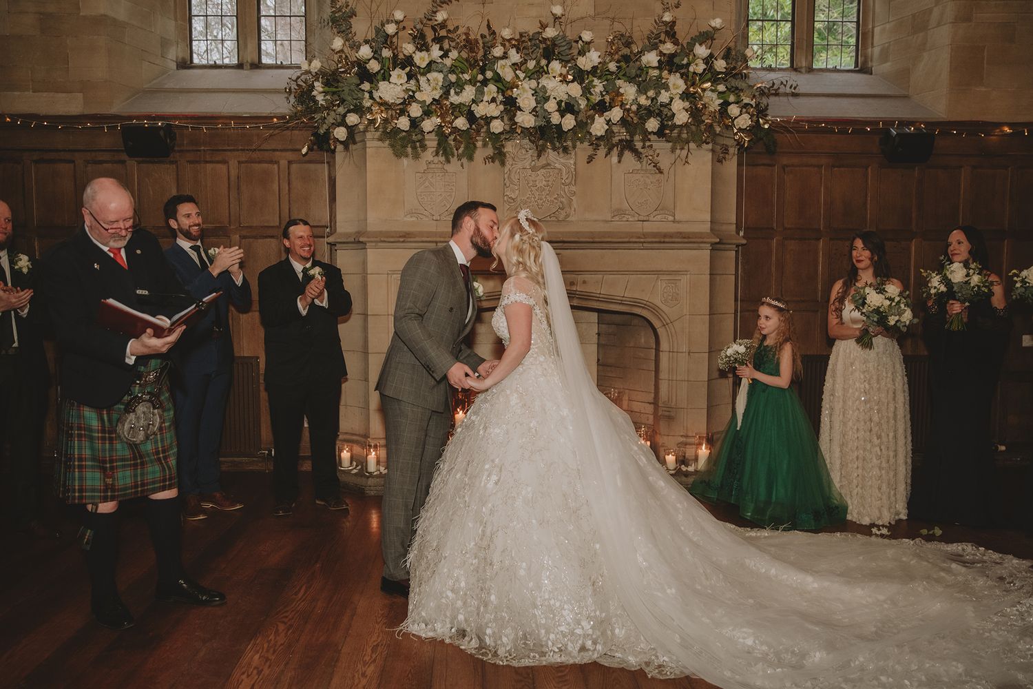 Bride and groom share first kiss at elegant castle wedding ceremony with floral arch and bridal party in green gowns.
