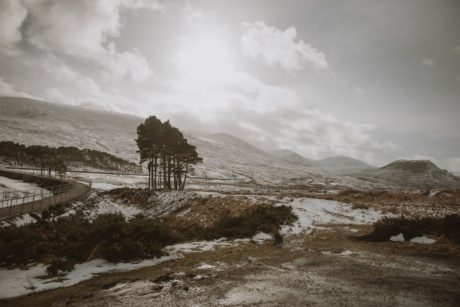 Snow-dusted highland landscape with lone trees, winding road, and dramatic cloudy sky over misty mountains.