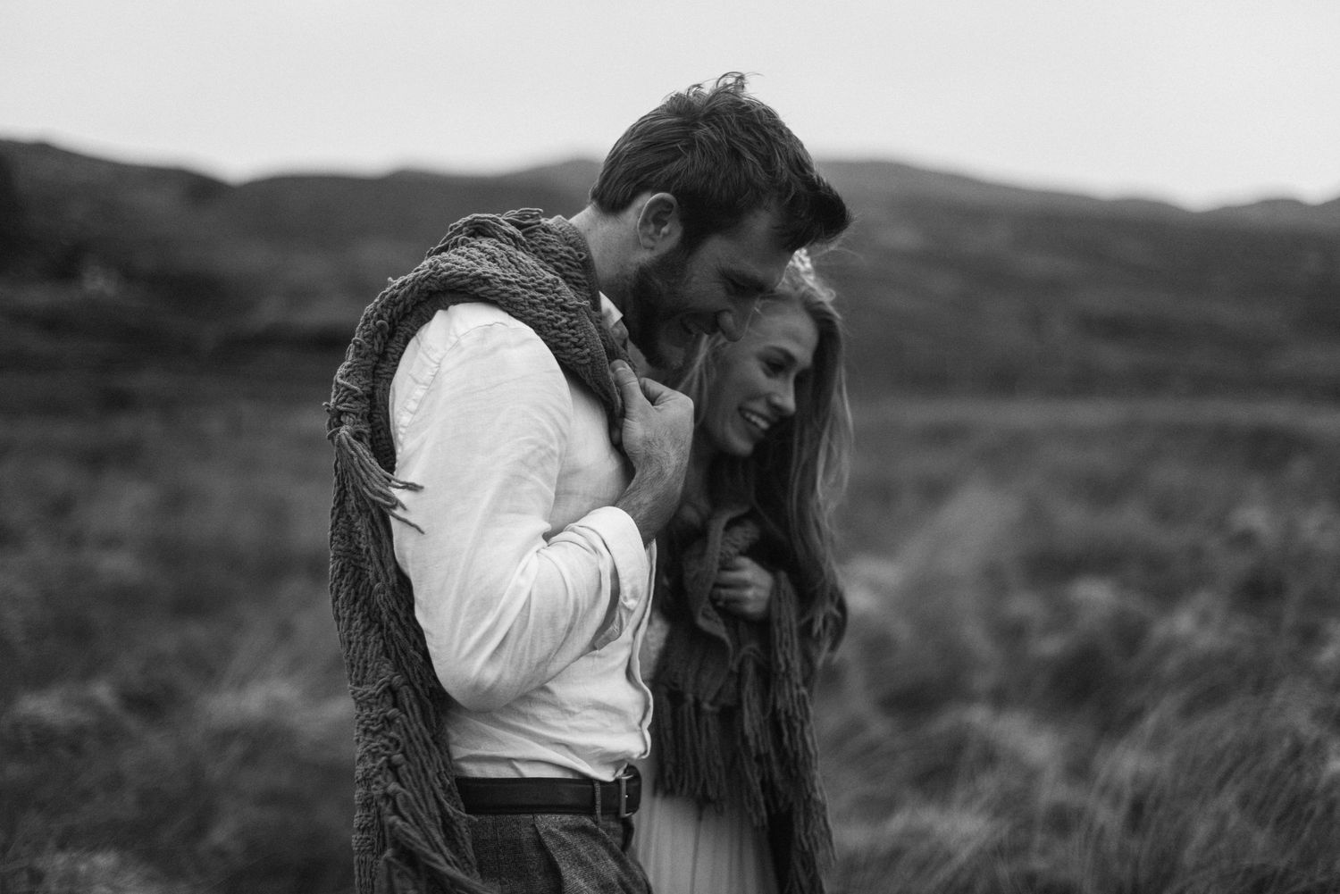 Black and white photo of couple wrapped in a blanket sharing a tender moment in a field.