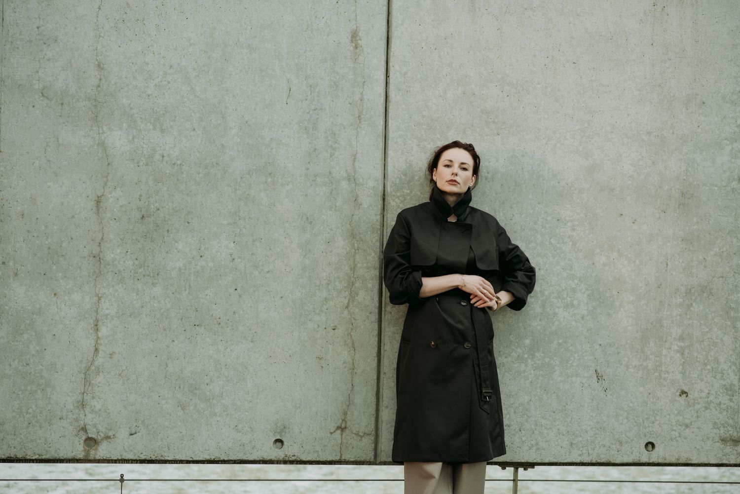 Elegant woman in dark green double-breasted trench coat stands against weathered concrete wall.