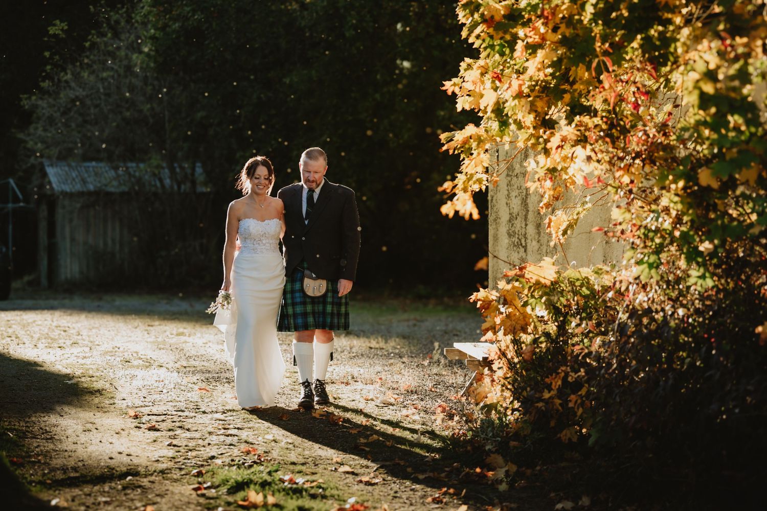 Bride and groom walk through autumn foliage in golden sunlight, groom wearing a traditional Scottish kilt.