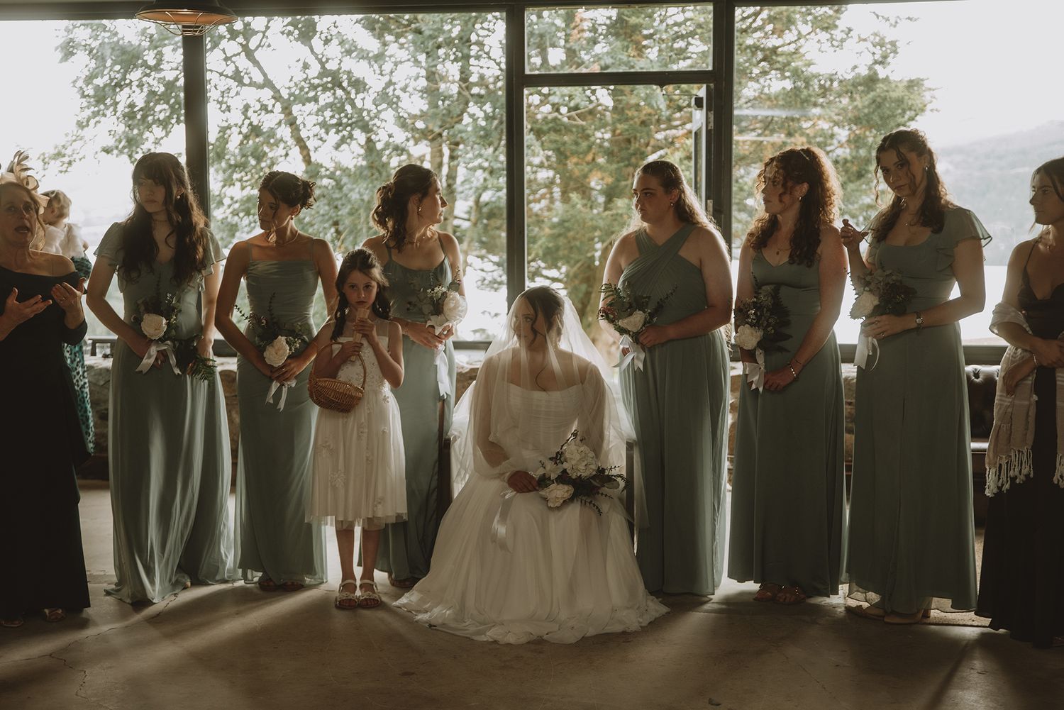 Bride seated with bridesmaids in sage green dresses standing by large window with scenic view.