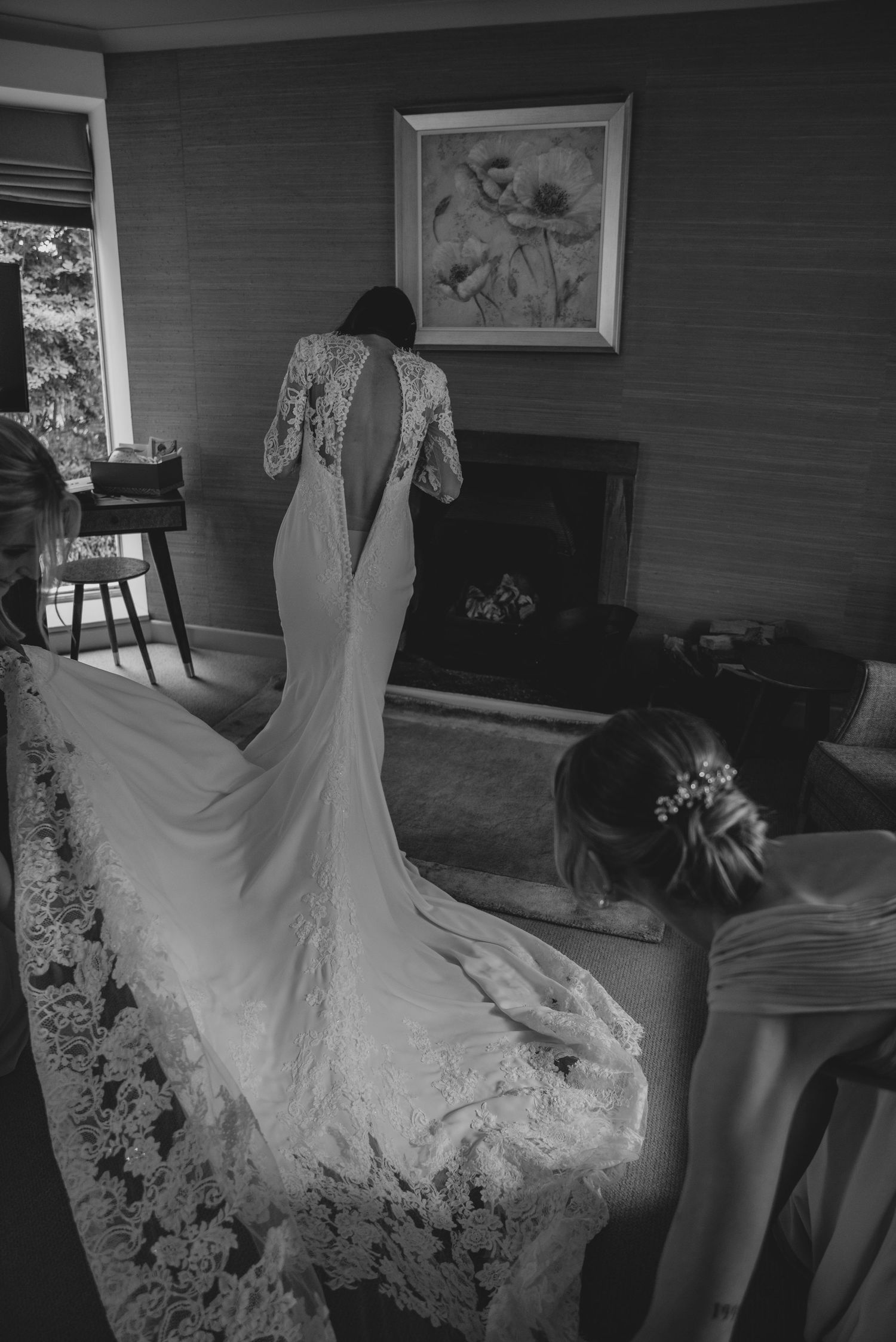Black and white photo of bride in lace back wedding gown with train, assisted by bridesmaid near fireplace.