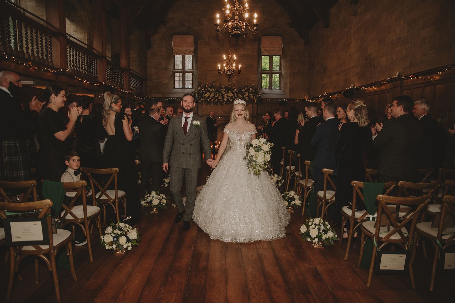 Bride and groom walk down the aisle of a grand hall, surrounded by cheering guests in elegant attire.