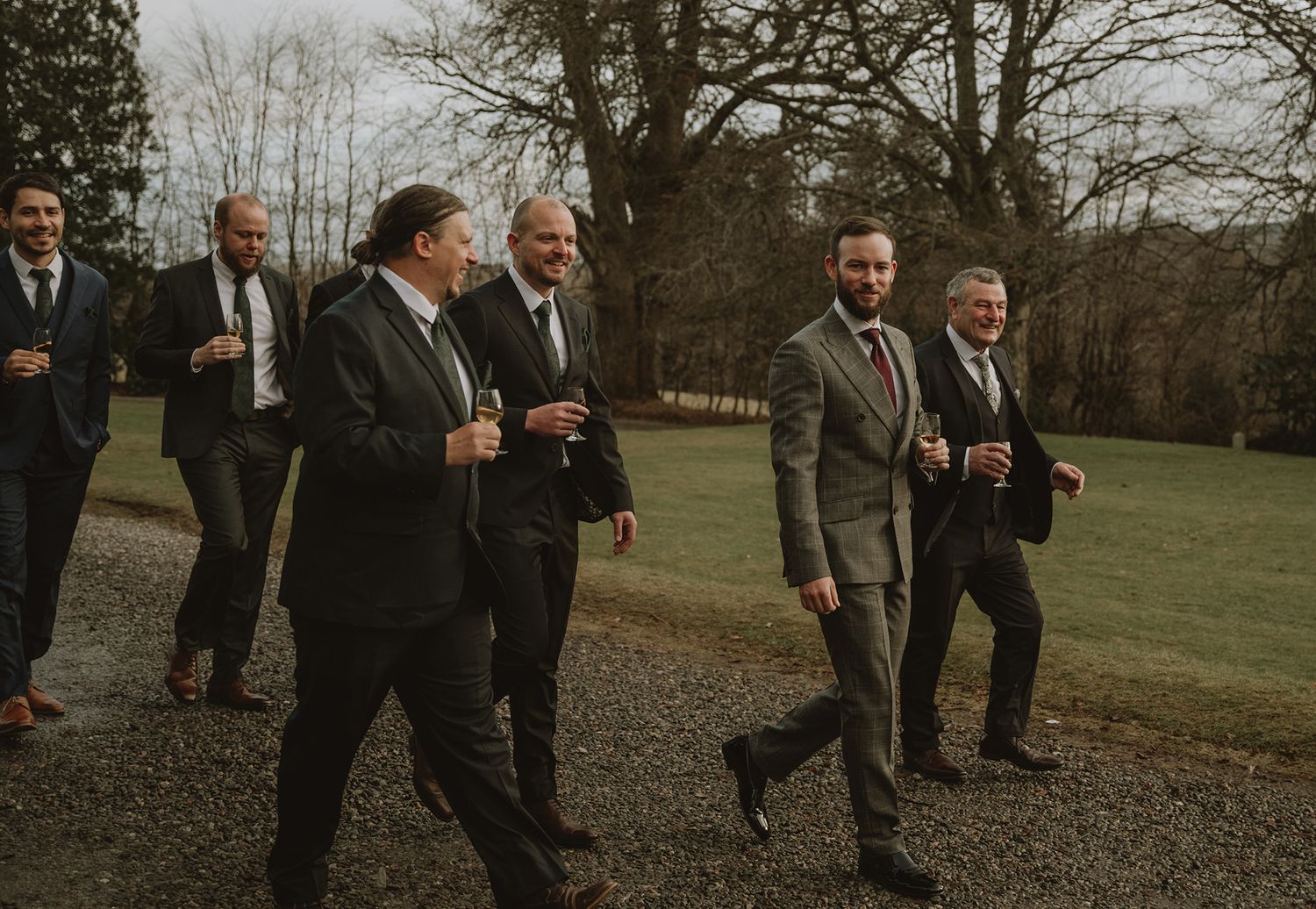 Groomsmen in dark suits laughing and walking outdoors at a wedding venue surrounded by green trees.
