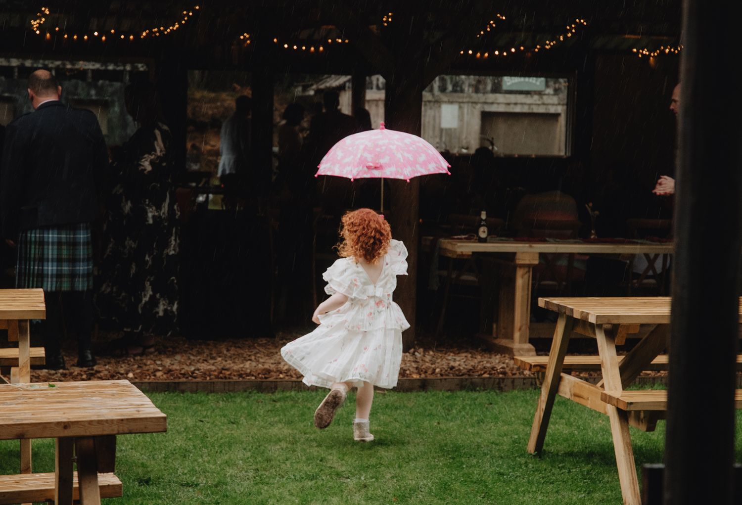 Young girl in white dress running with pink umbrella on grass at outdoor wedding venue.