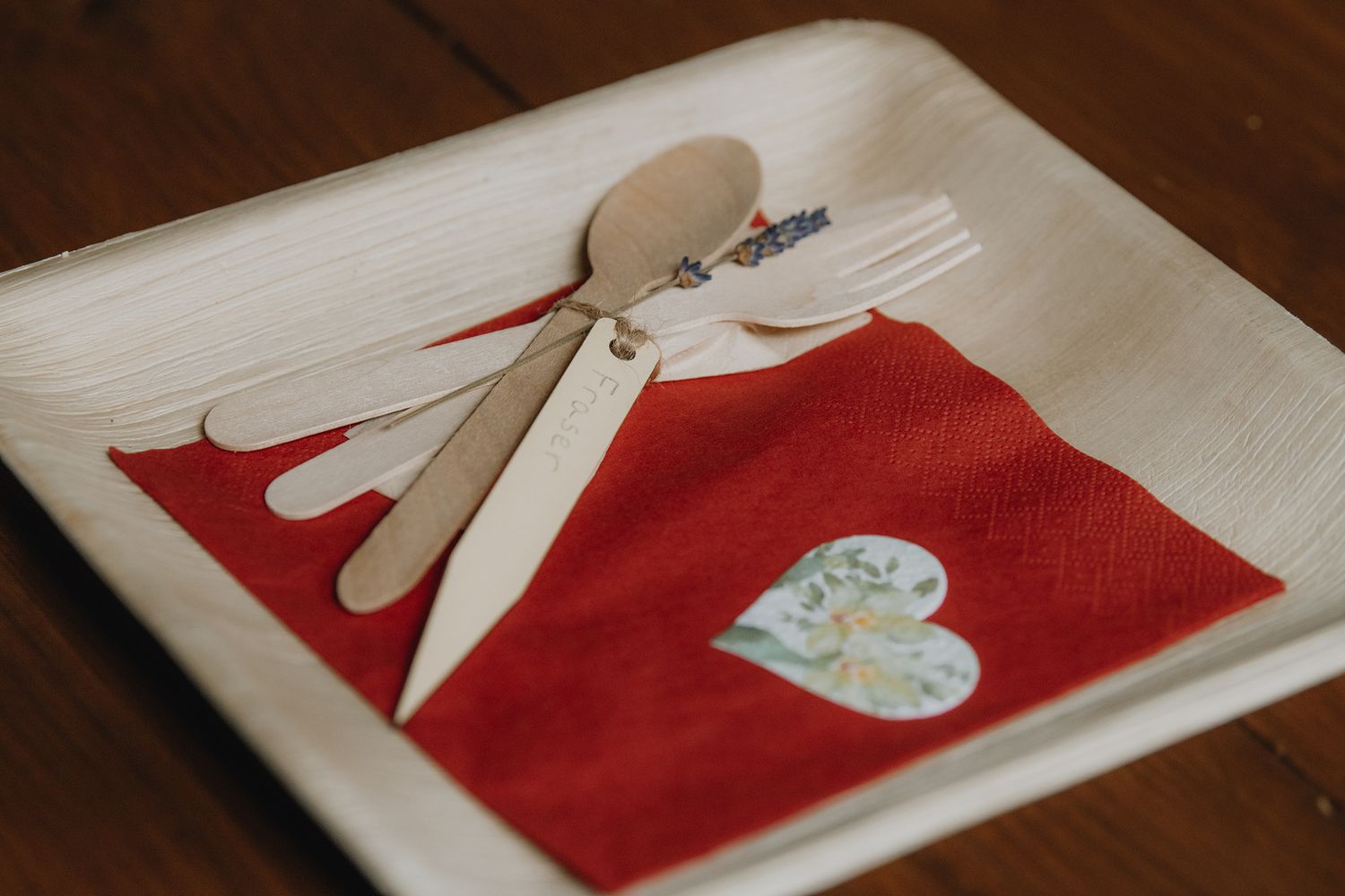 A rustic wedding place setting with wooden cutlery and a red napkin on a palm leaf plate.