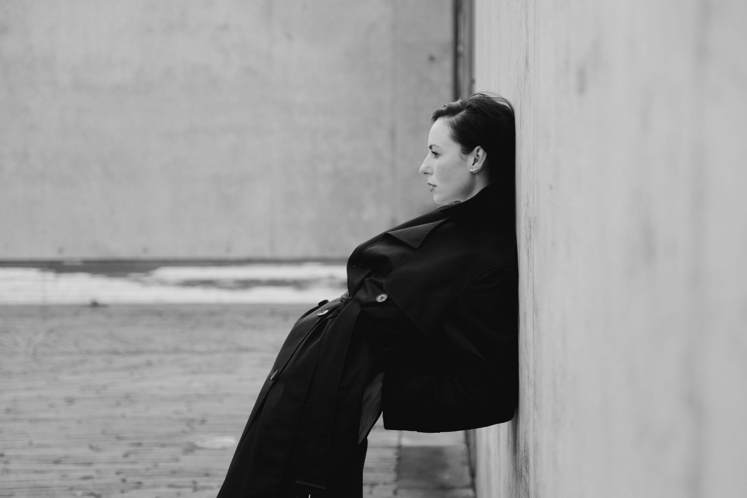 Black and white photo of a woman in a dark coat leaning against a concrete wall, gazing thoughtfully into the distance.