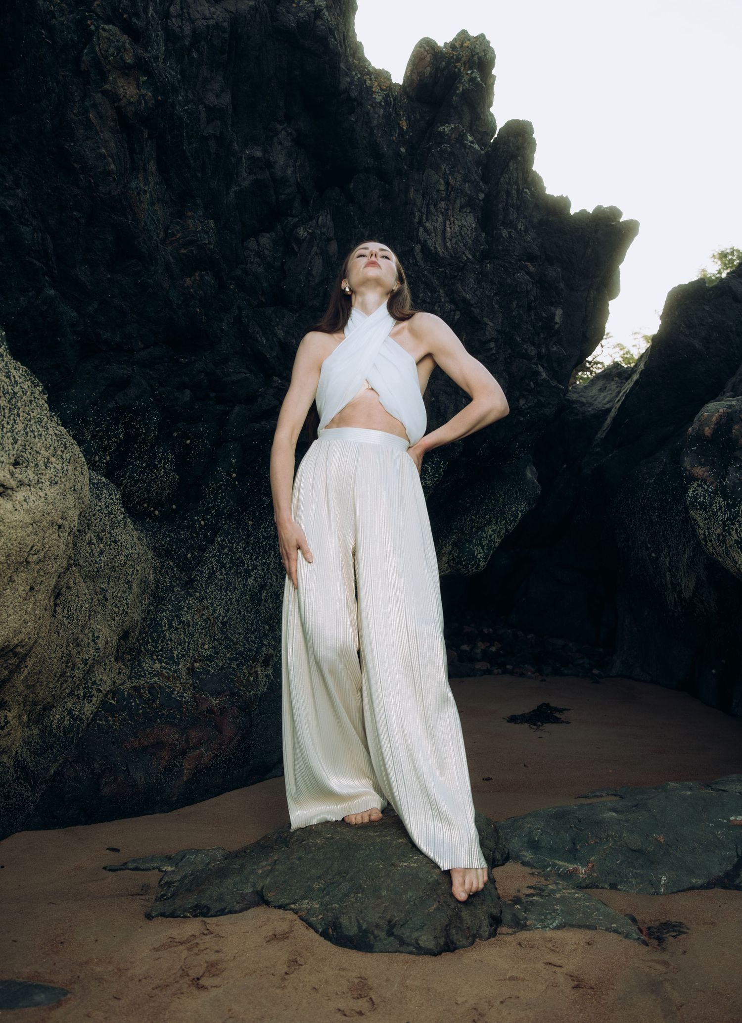 Woman in white halter crop top and wide-leg trousers posing among dark coastal rocks on sandy beach.