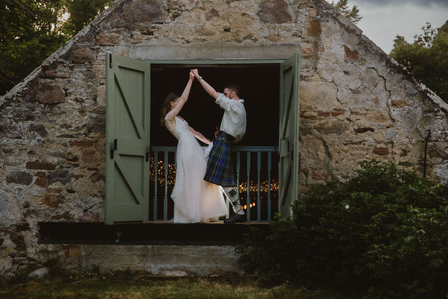 Bride and groom dancing in doorway of rustic stone barn decorated with fairy lights at dusk.