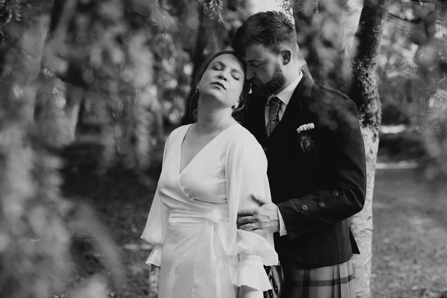 Black and white intimate photo of groom embracing bride from behind among blossoming trees.