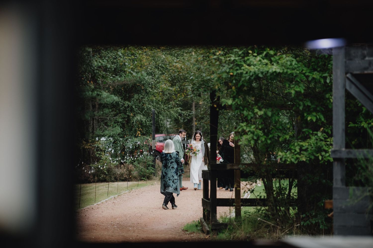 Wedding party gathered at a wooden gate entrance surrounded by lush green forest trees.