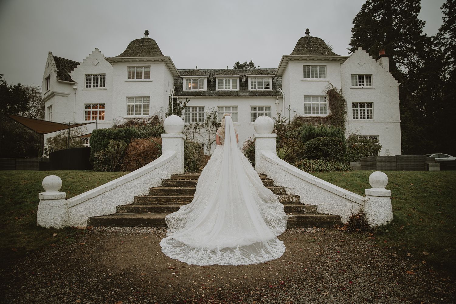 Bride in flowing white gown with long train poses on grand staircase of elegant white manor house.