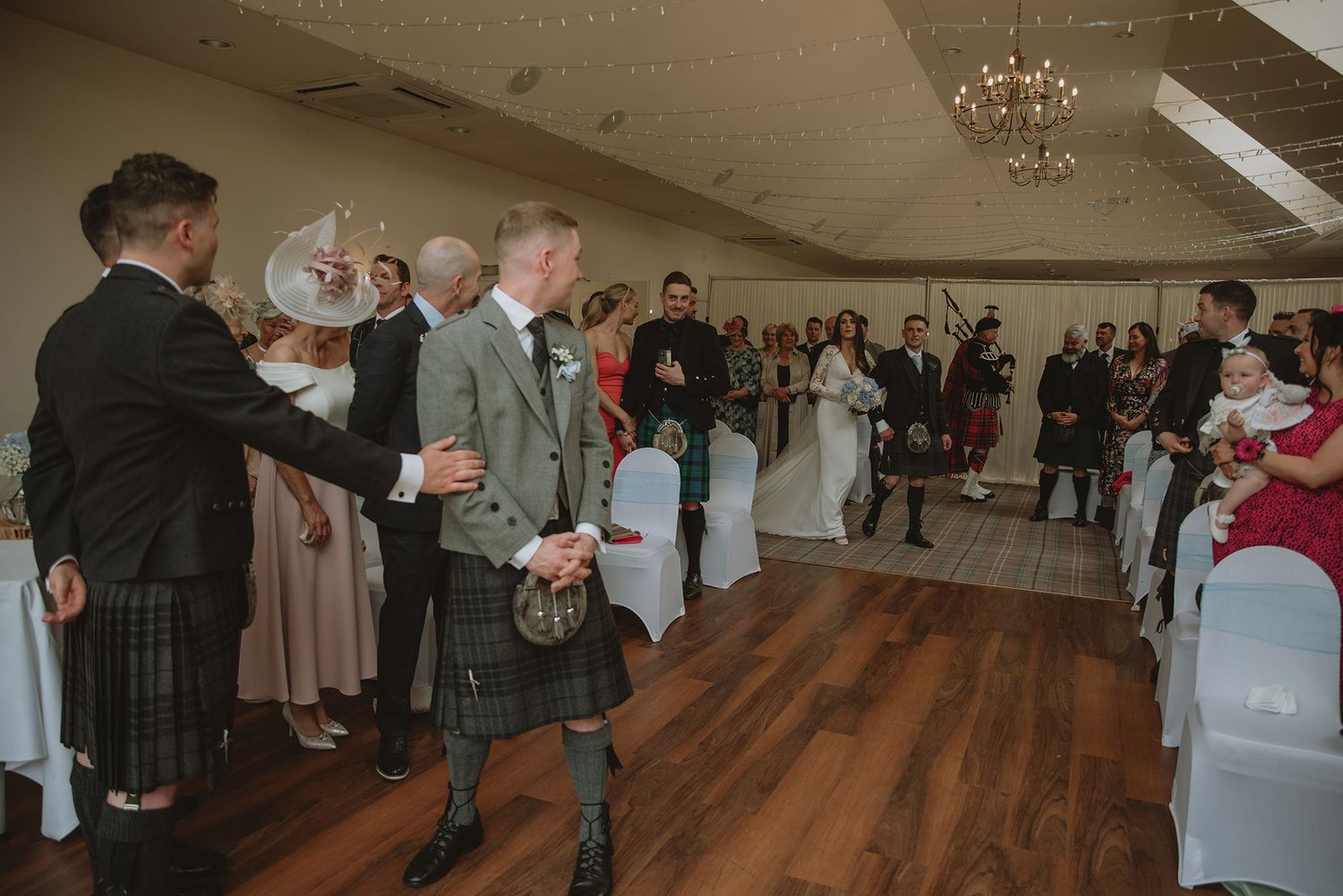 Scottish wedding ceremony with guests in kilts and formal attire gathered in an elegant venue with wooden floors.