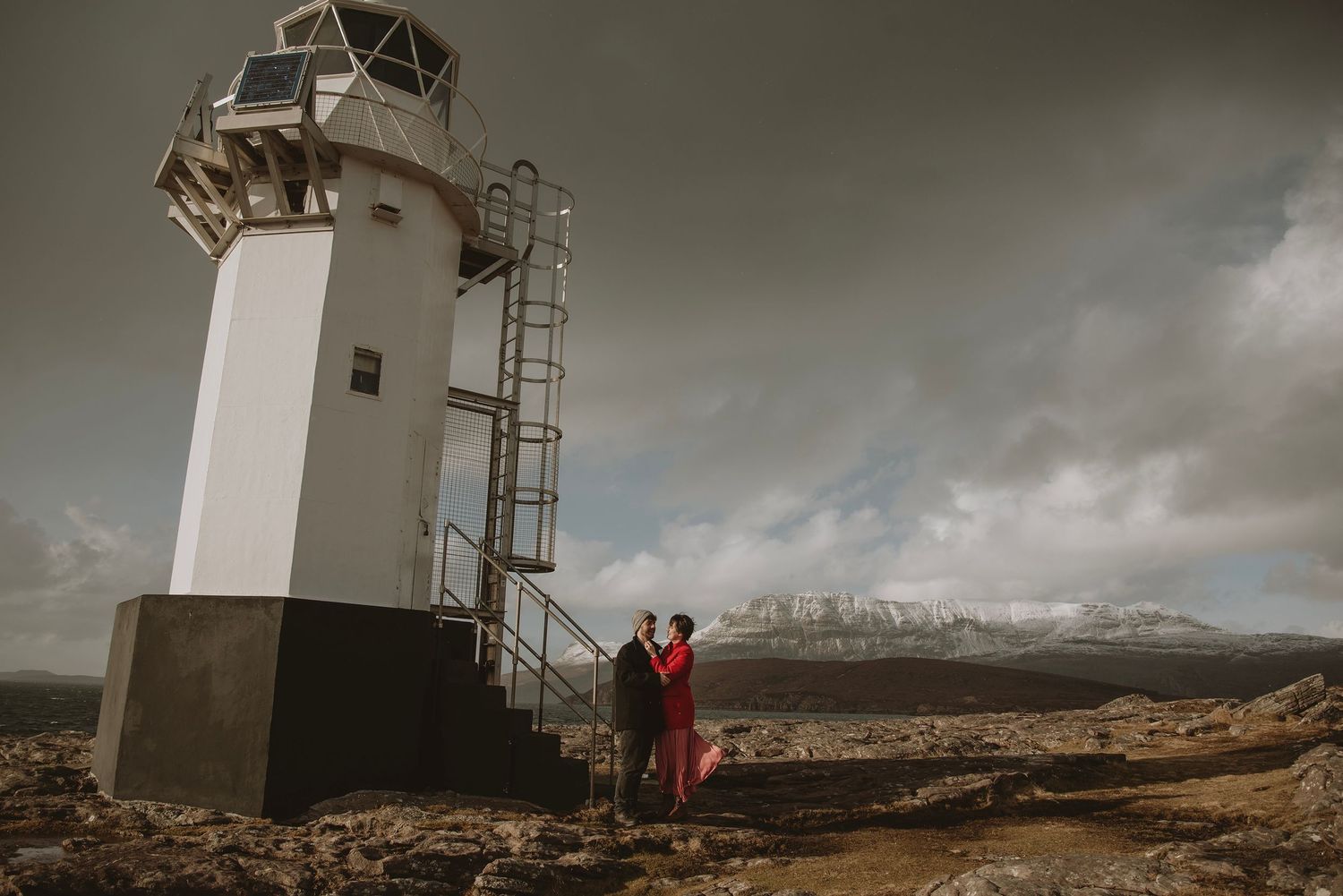 Couple embracing near a white lighthouse on rocky terrain under dramatic stormy skies in Iceland.