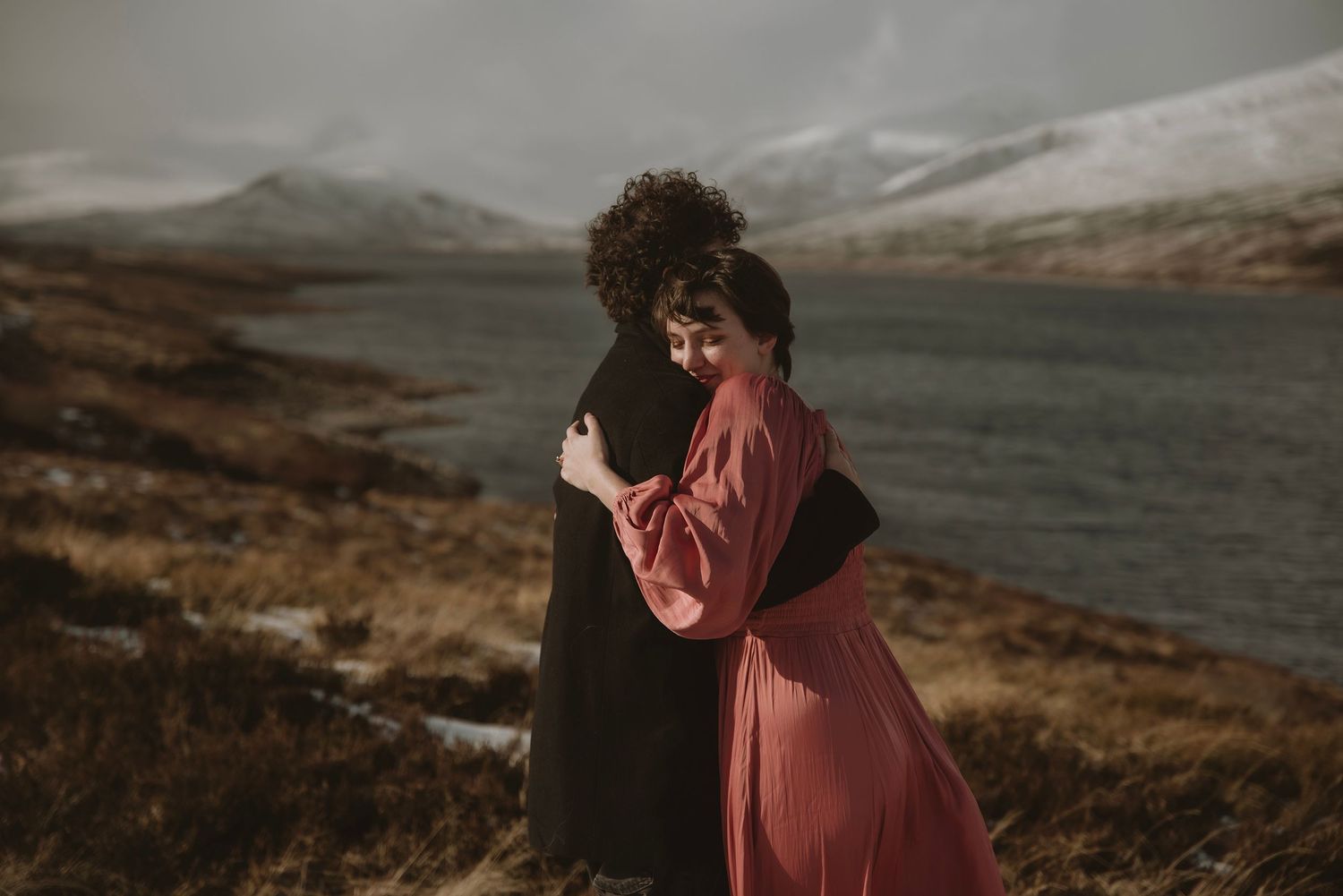 Couple embracing romantically by a scenic loch with snow-capped mountains in the Scottish Highlands.