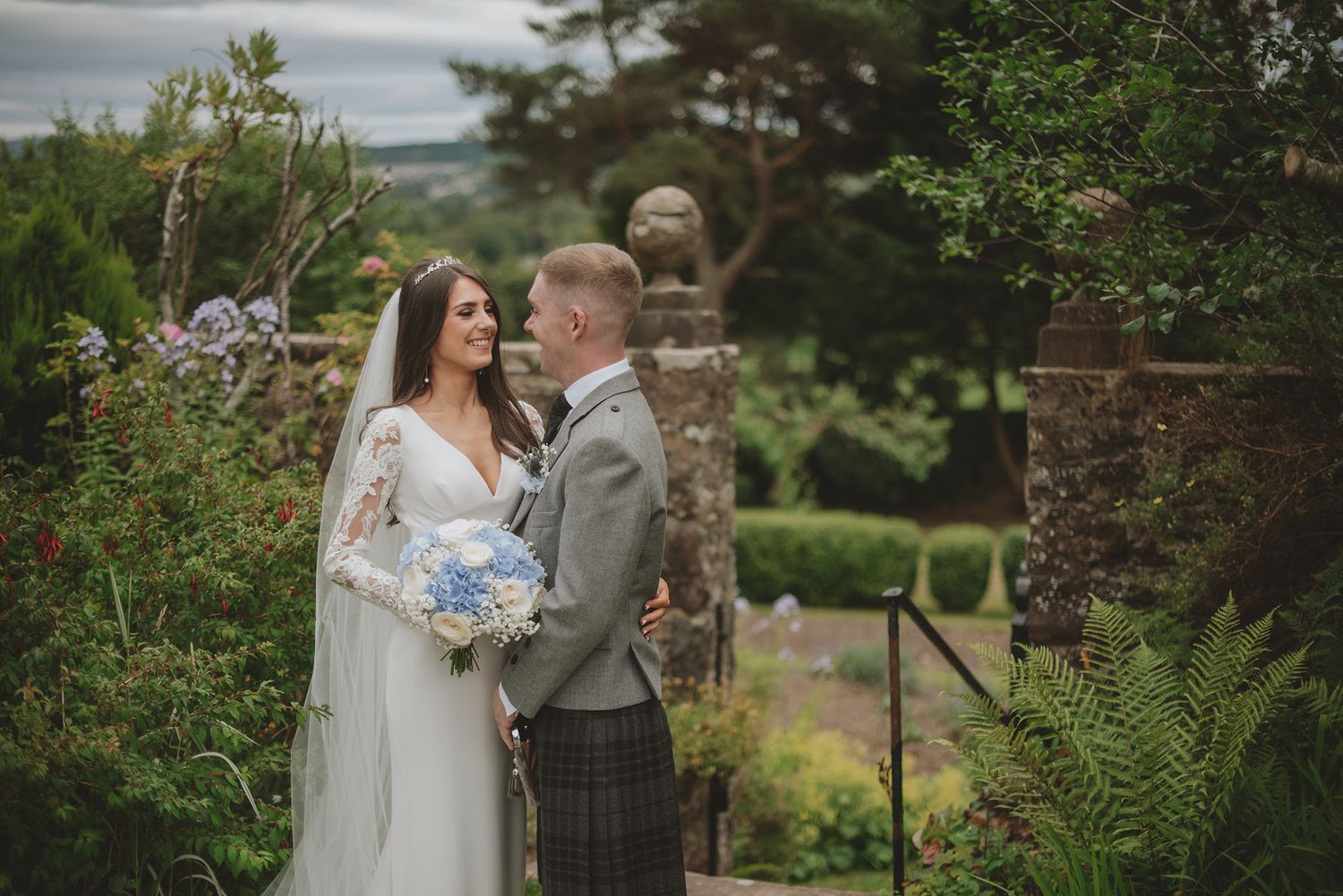 Bride and groom share a romantic moment in a lush garden, she in a white gown with veil, he in a traditional kilt.