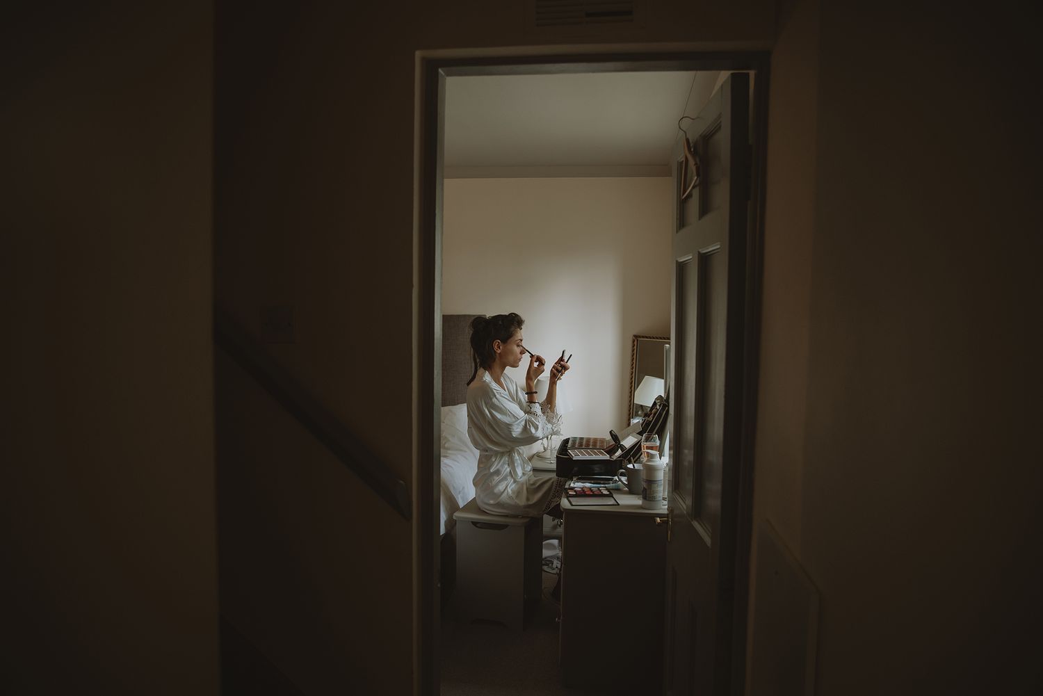 Bride in white robe applying makeup at vanity mirror, photographed through doorway in soft moody lighting.