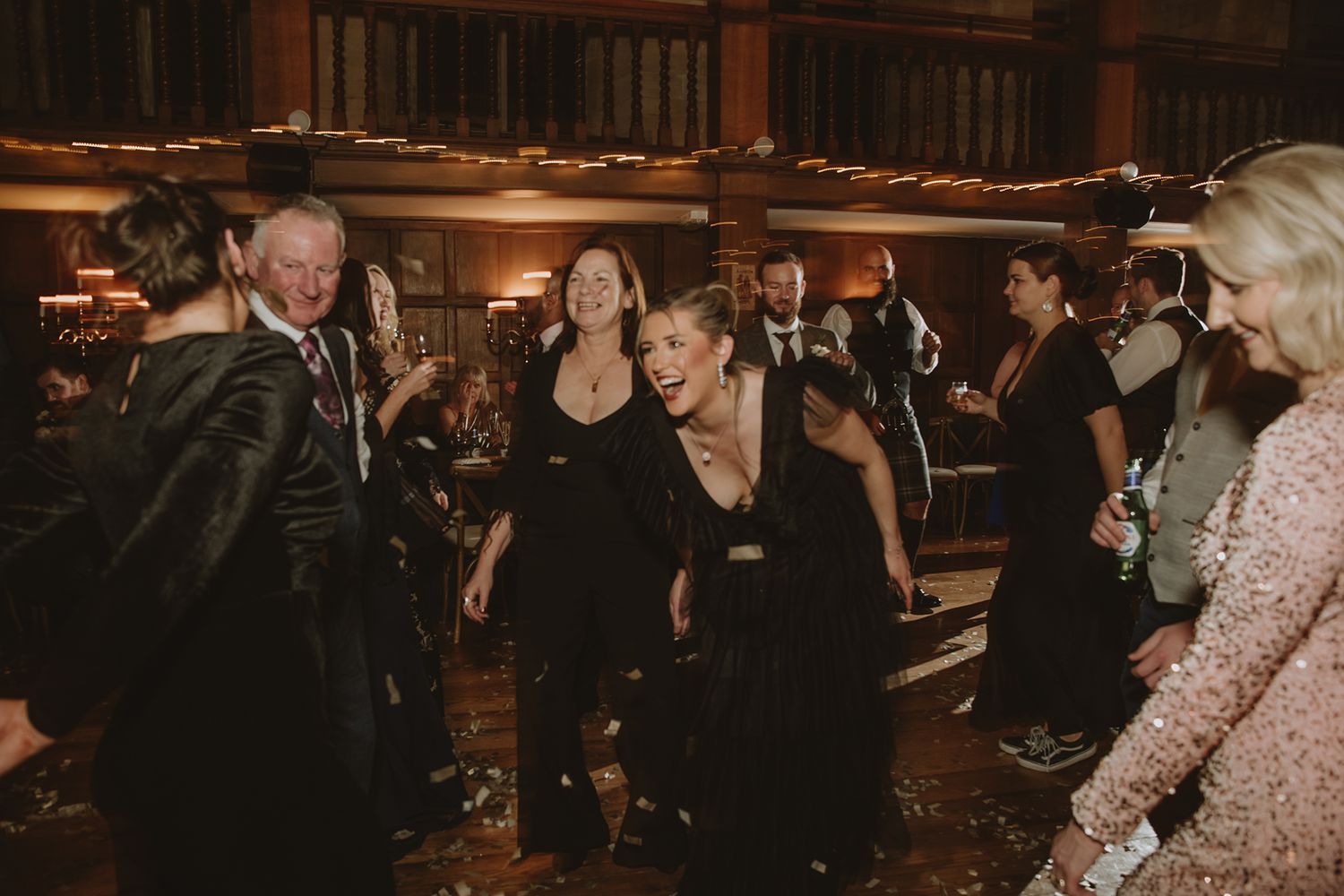 Guests in elegant black attire laugh and dance together at a glamorous evening event in a warmly lit venue.