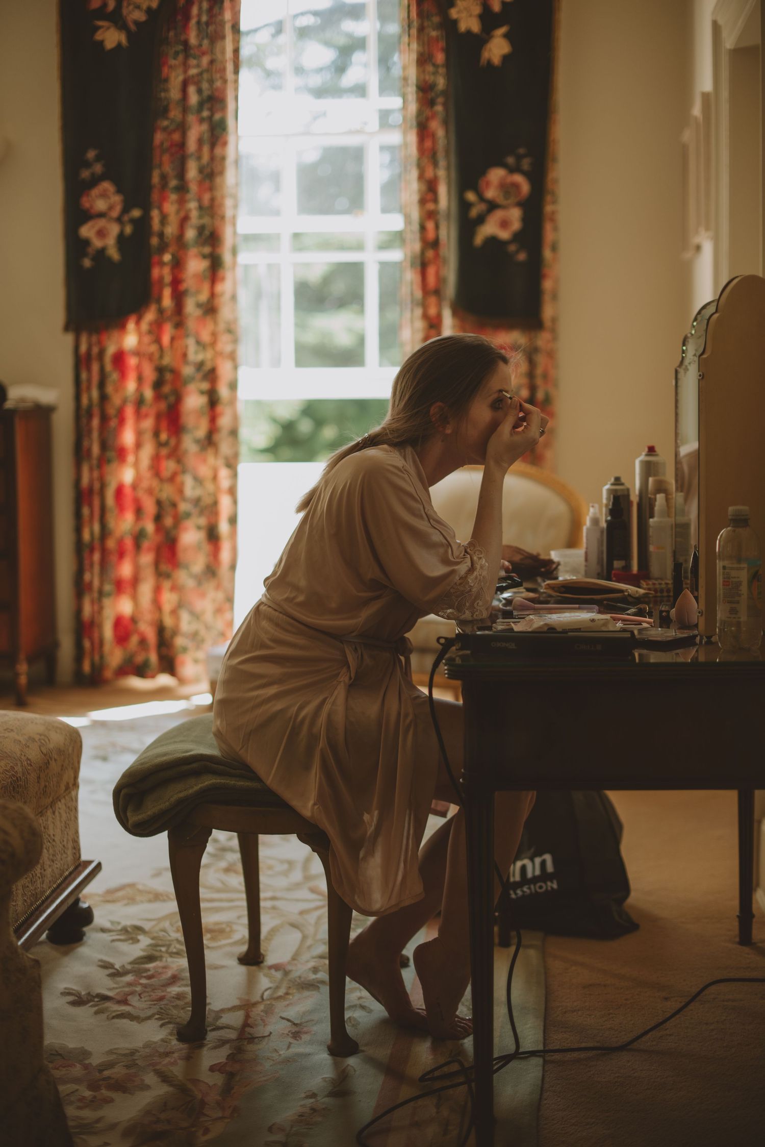 Bride in silk robe applies makeup at vintage vanity table before her wedding ceremony.