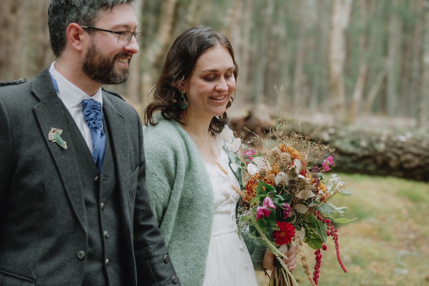 Smiling bride holding vibrant autumn bouquet beside bearded groom in green tartan jacket in forest.