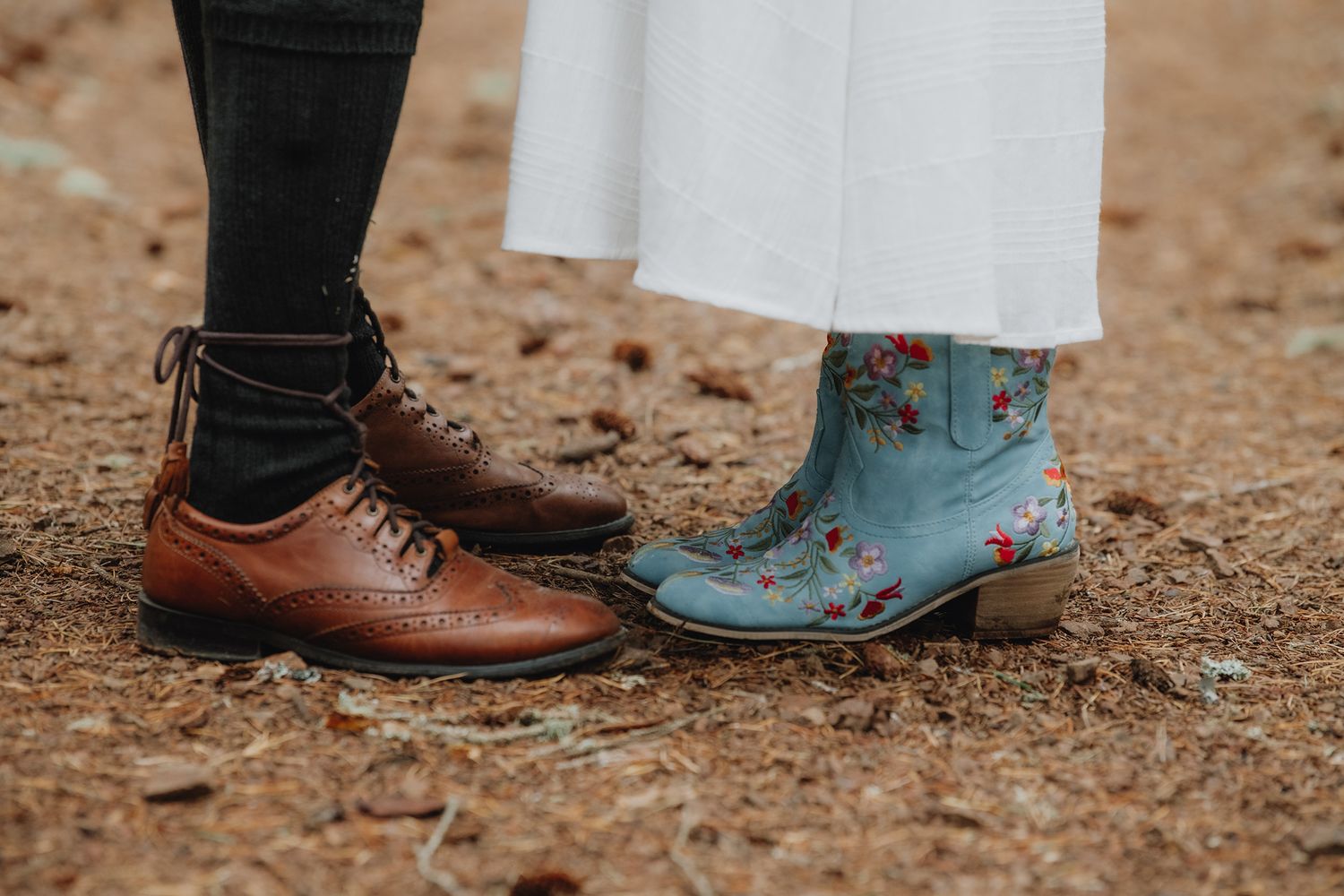 Side-by-side shot of groom's brown brogue shoes and bride's floral blue cowboy boots on sandy ground.