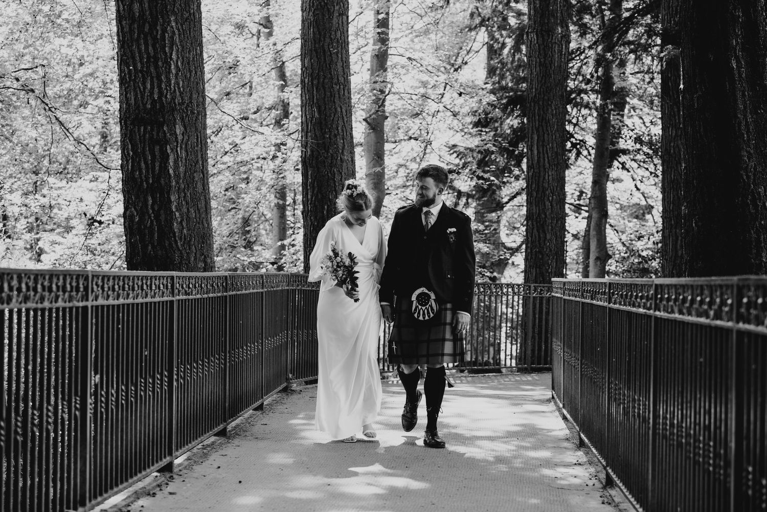 Black and white photo of bride and groom walking across ornate bridge through woodland setting.