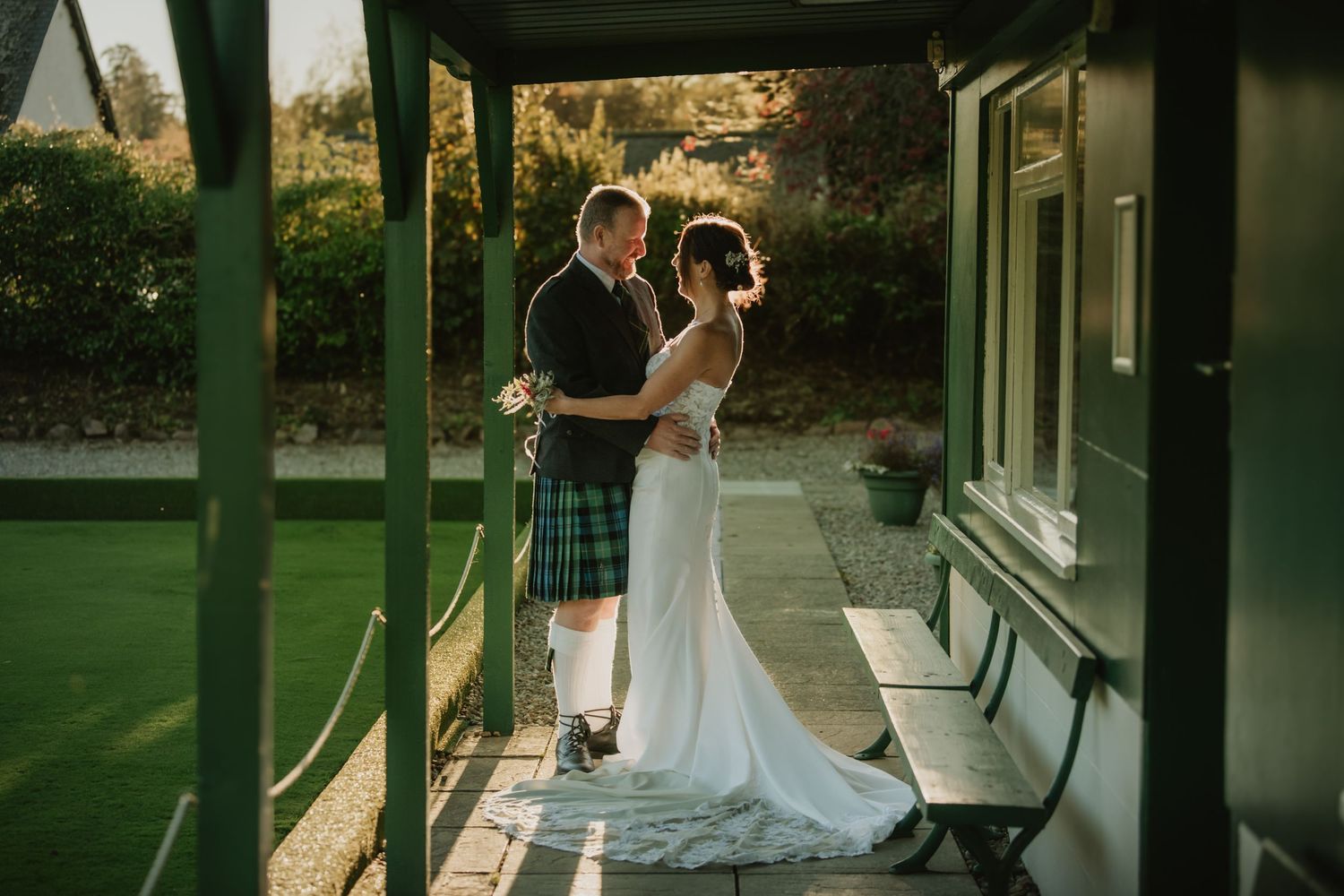 Bride in white gown and kilted groom share a tender embrace on a sunlit covered porch.