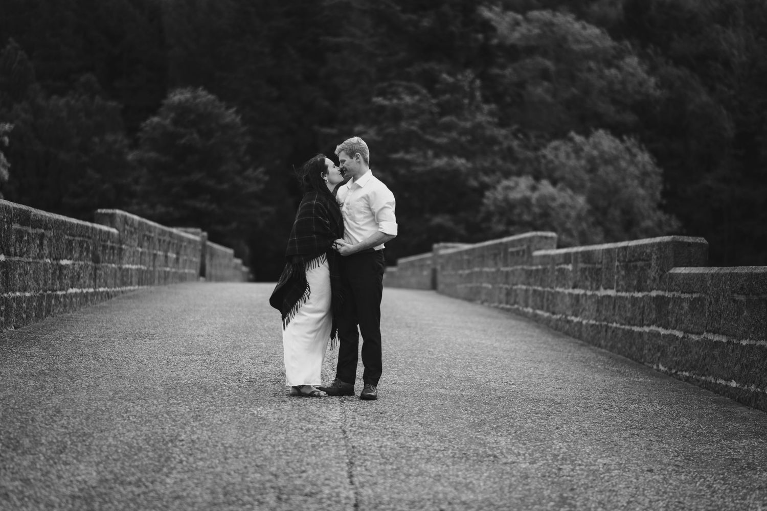 Couple sharing a tender kiss on a stone bridge surrounded by lush green trees.