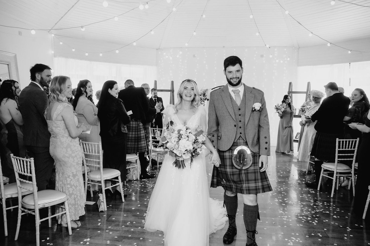 Newlyweds walk down aisle together as groom wears traditional Scottish kilt in black and white photo.