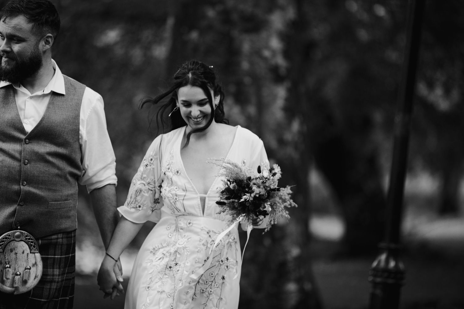 Black and white photo of laughing bride holding colorful bouquet walking with kilted groom outdoors.