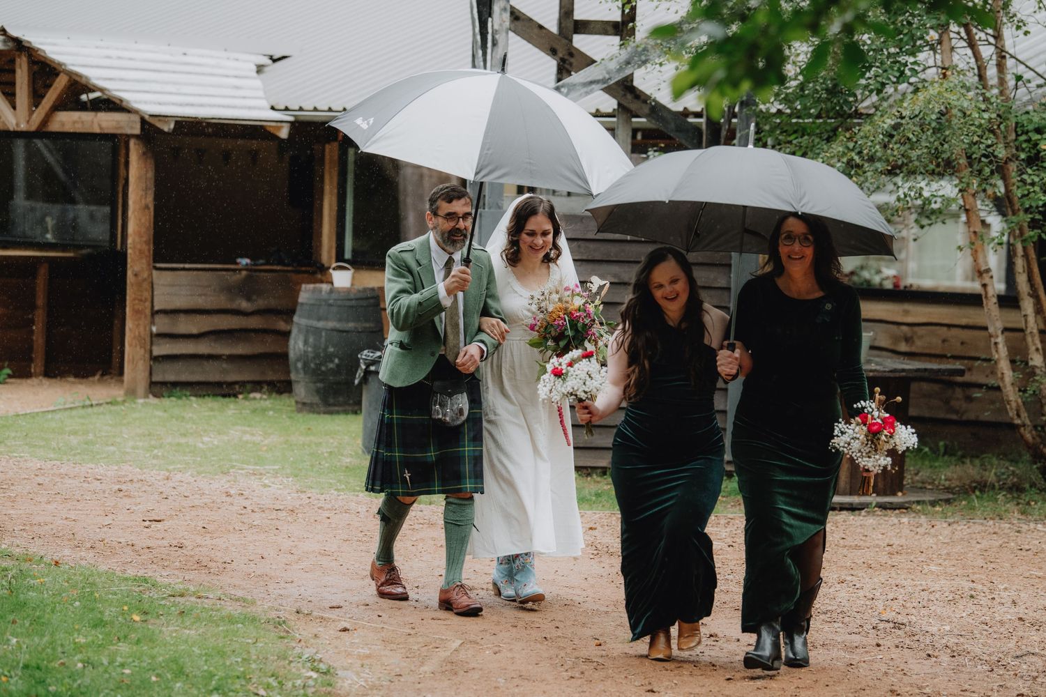 Bride and groom in kilt walk with guests under umbrellas on rainy wedding day.