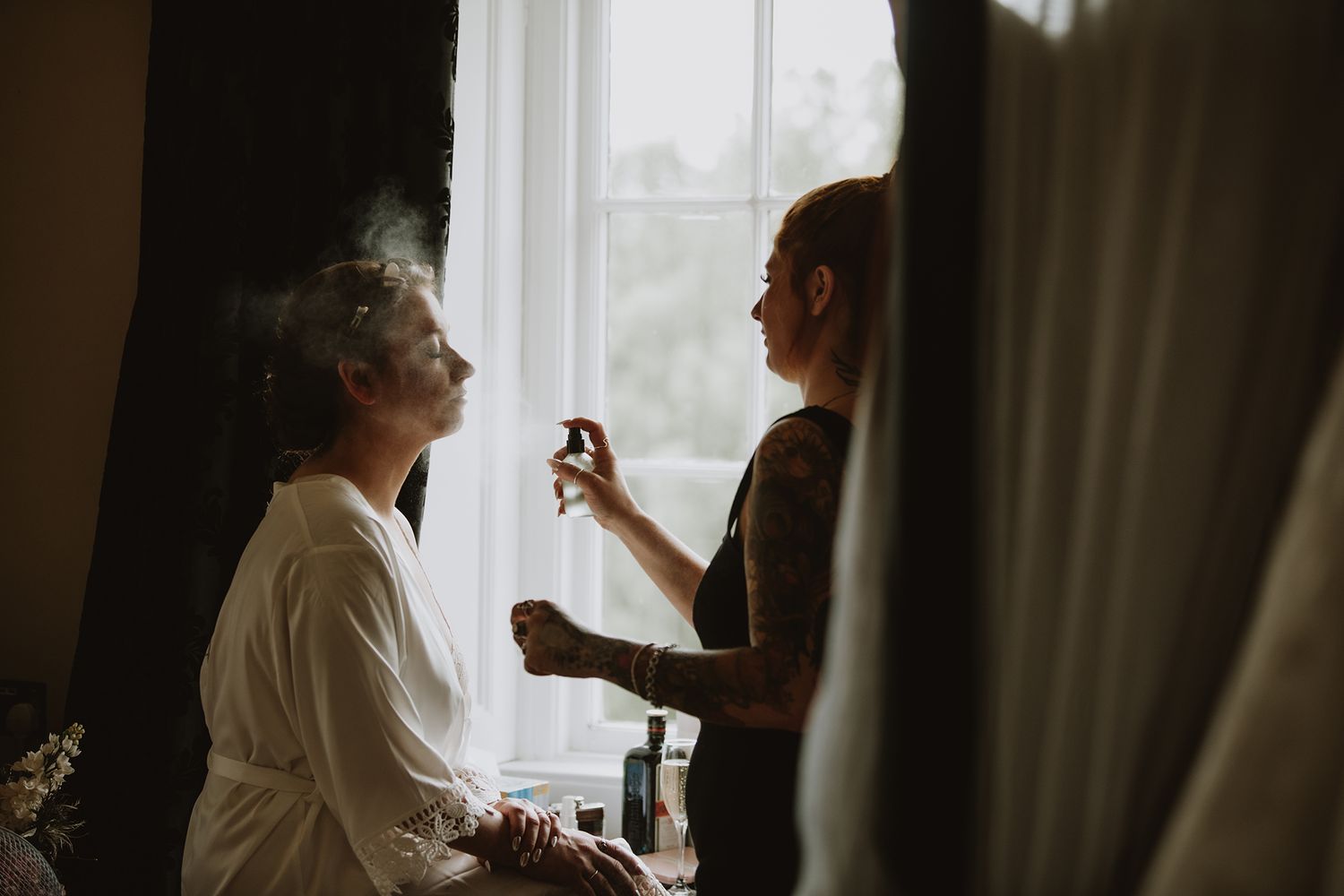 Bridesmaid applying setting spray to bride seated by window during wedding morning prep.