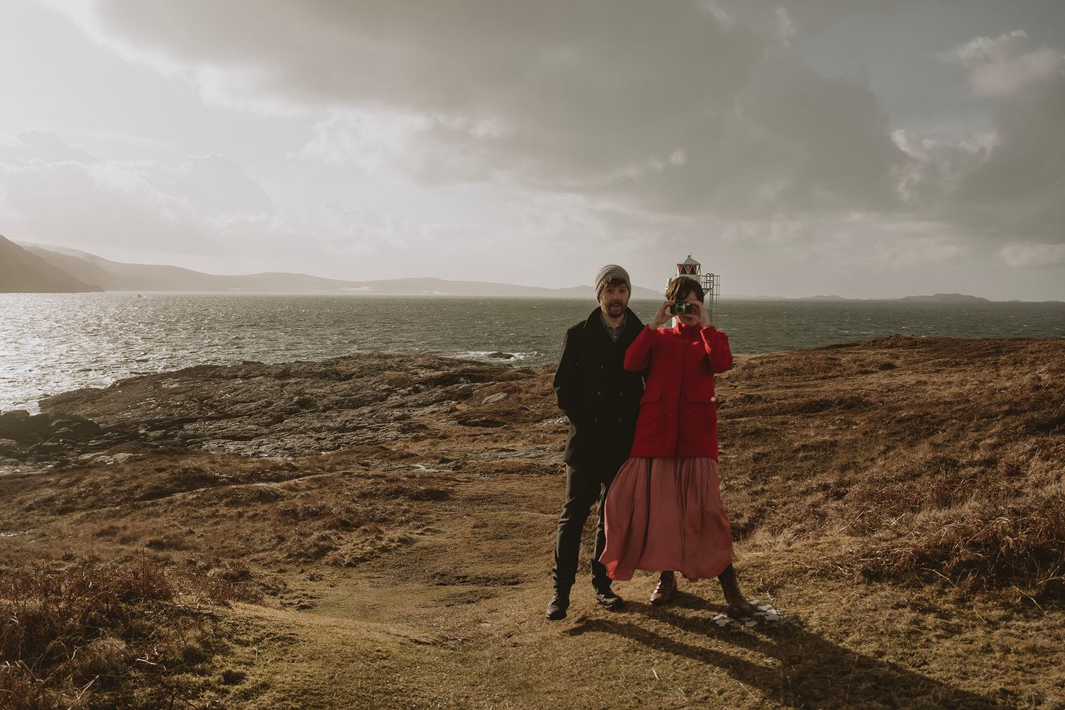Couple standing on coastal hillside, woman in red dress, overlooking calm waters under golden cloudy sky.