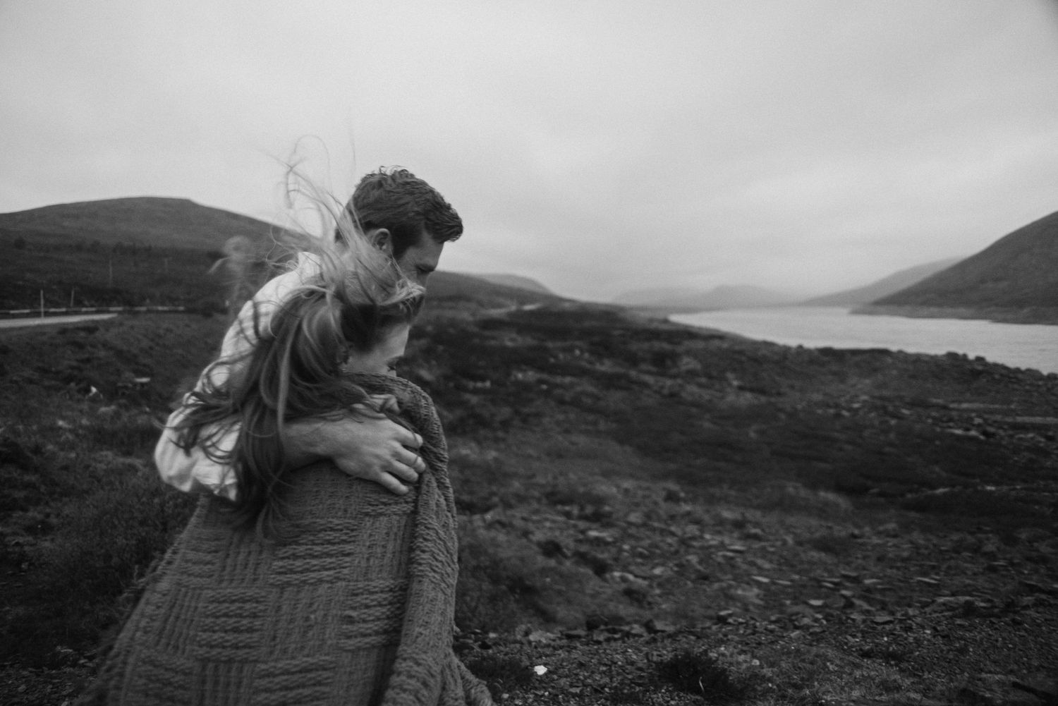 Black and white photo of a couple embracing near a scenic loch with misty hills.