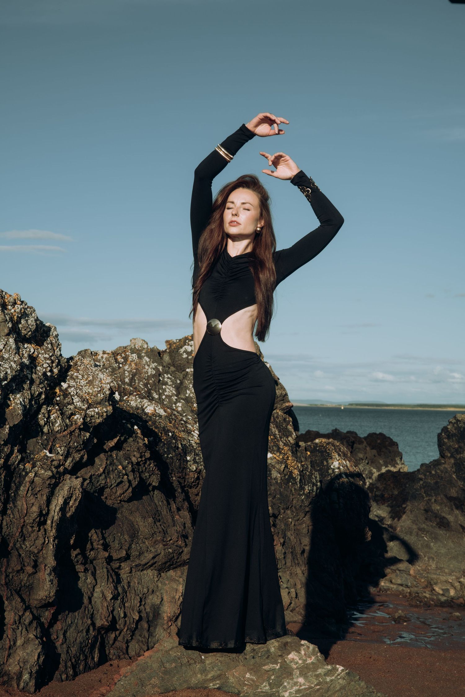 Woman in black cutout gown posing gracefully on coastal rocks with ocean backdrop.
