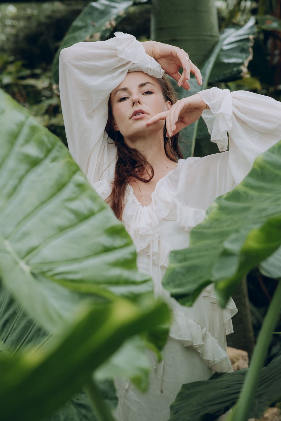 Woman in white ruffled bell-sleeve dress posing gracefully among large tropical leaves in a greenhouse.