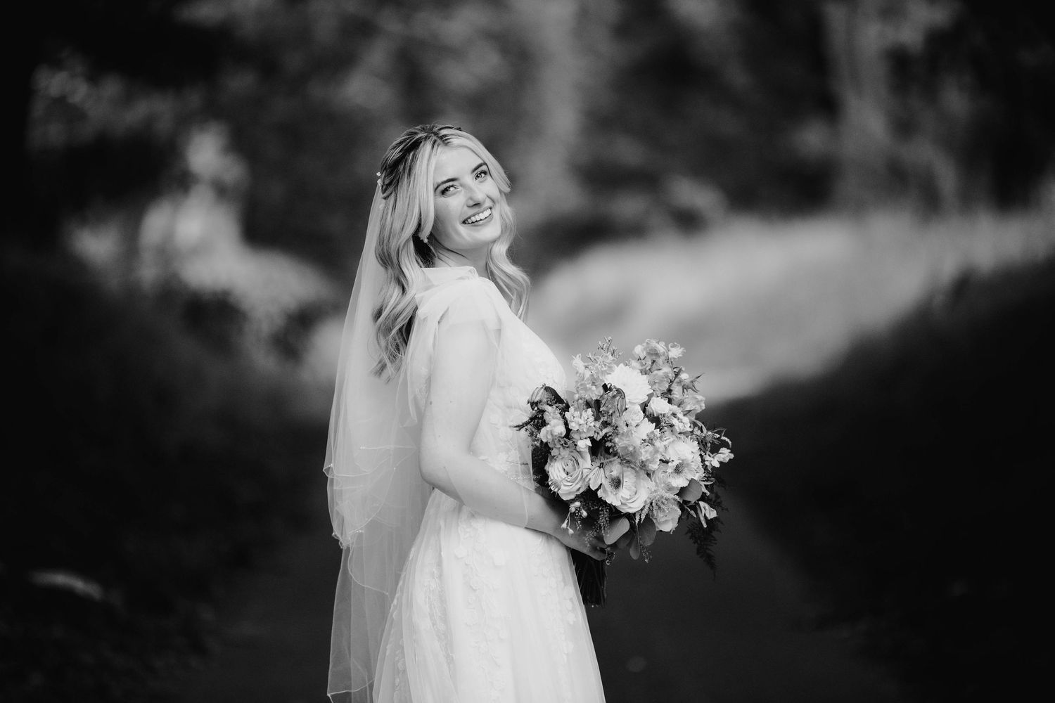 Smiling bride in white gown holding bouquet poses outdoors in black and white wedding portrait.