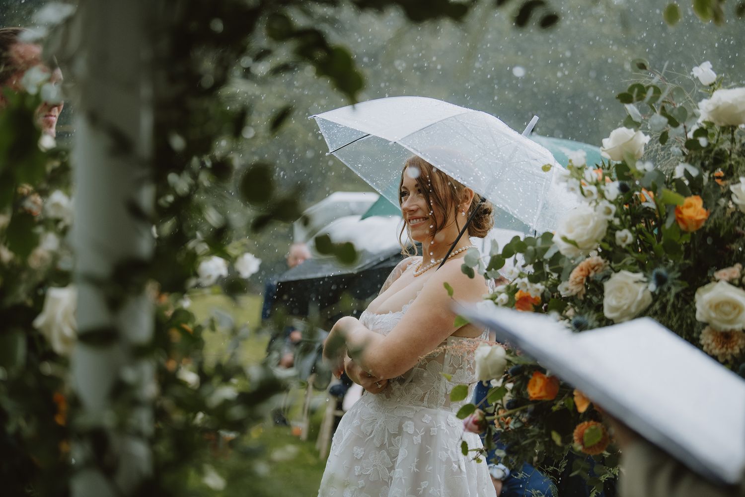 Bride in lace gown holding clear umbrella amid colorful floral arch in the rain.