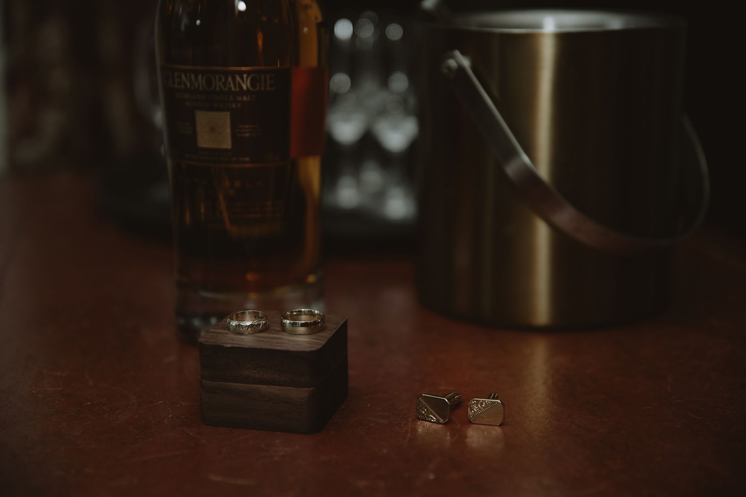 Wedding rings and cufflinks displayed beside a whiskey bottle and ice bucket on a wooden bar surface.