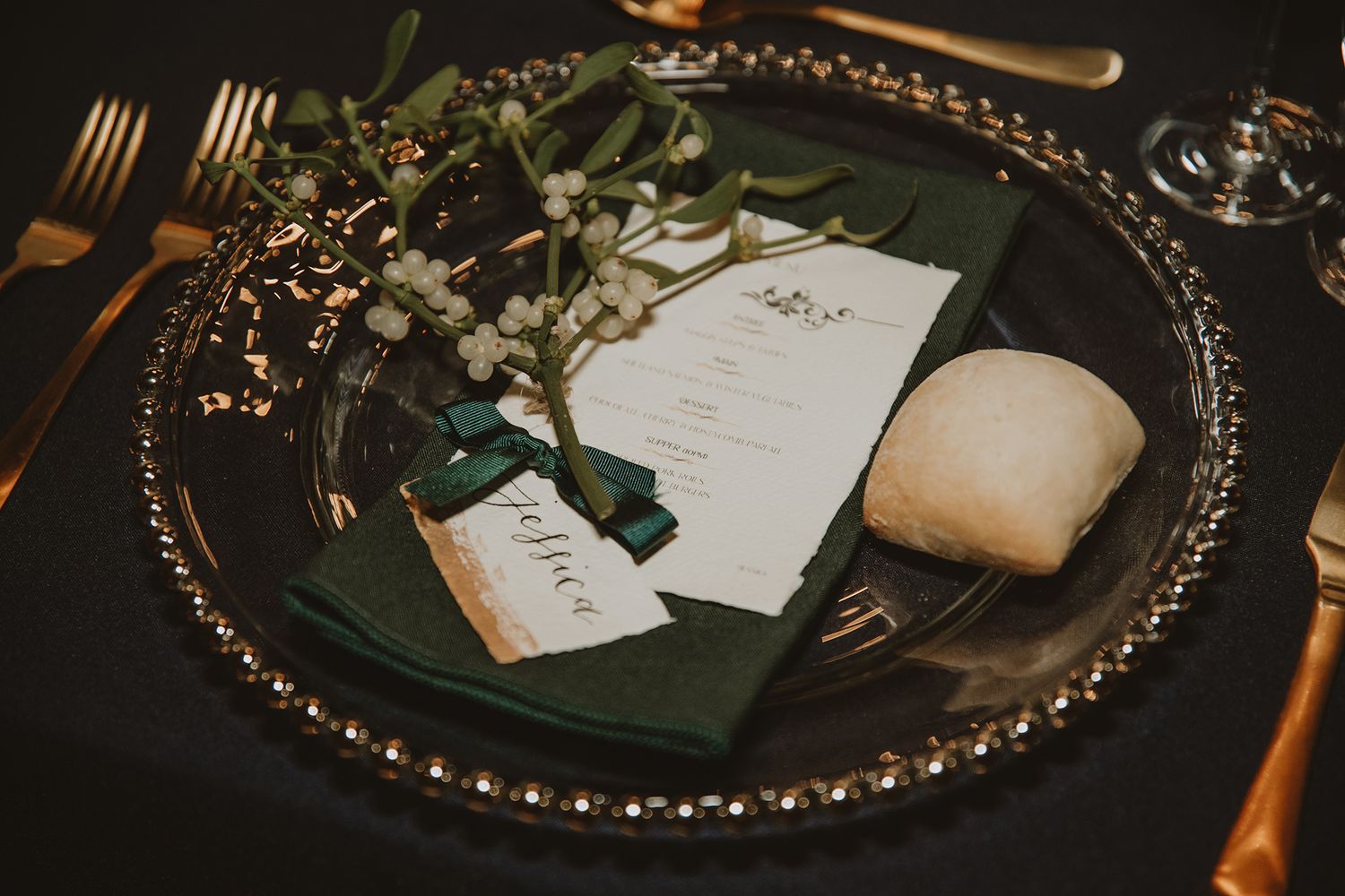 Elegant wedding place setting with green napkin, floral sprig, menu card, and gold cutlery on a silver charger plate.