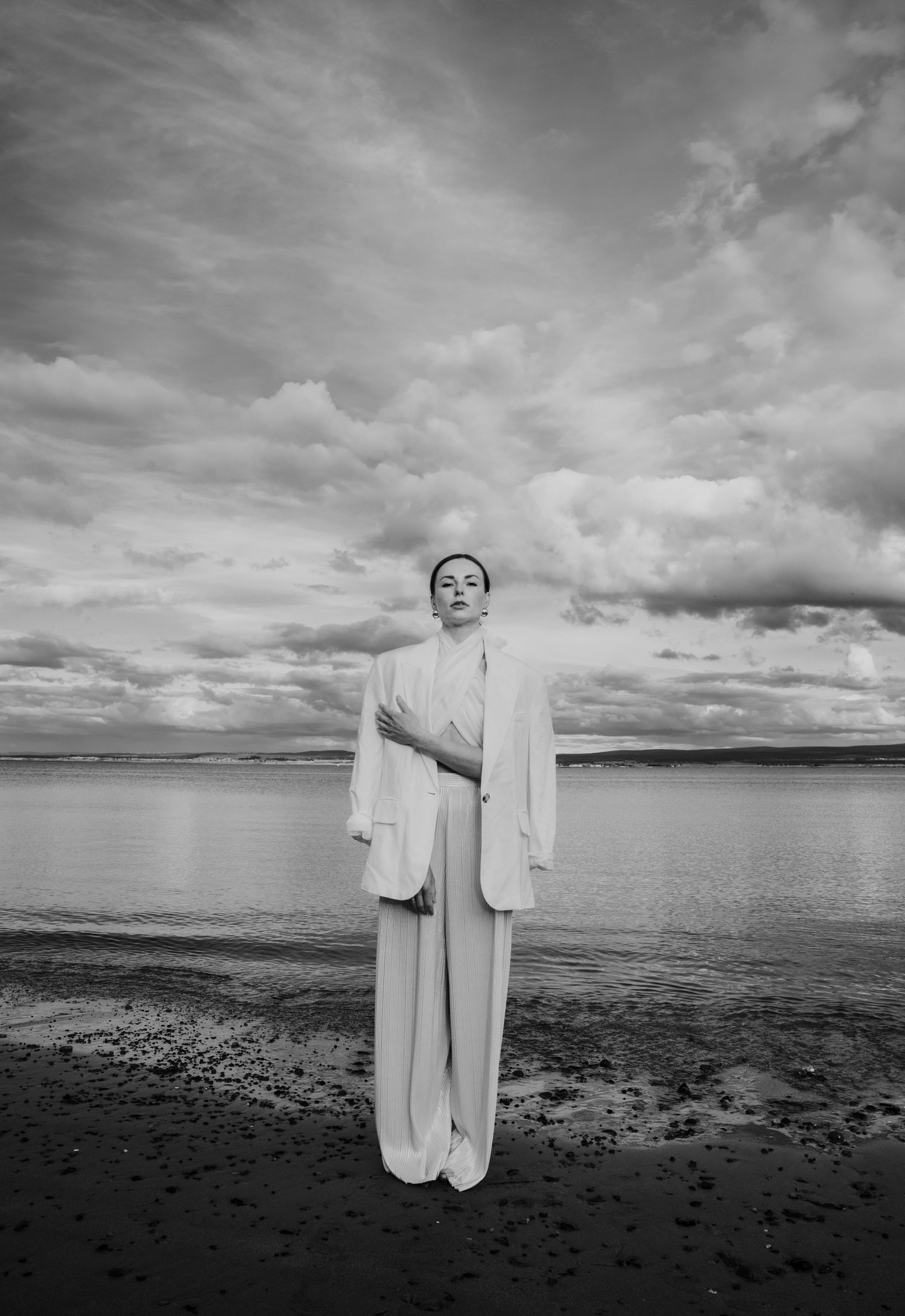Black and white photo of woman in white blazer and wide-leg pants standing on rocky shoreline under dramatic cloudy sky.