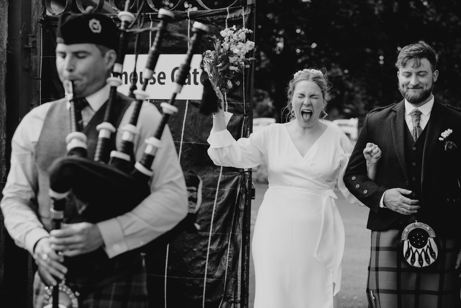 Black and white photo of joyful bride celebrating beside bagpiper and smiling groom at wedding.