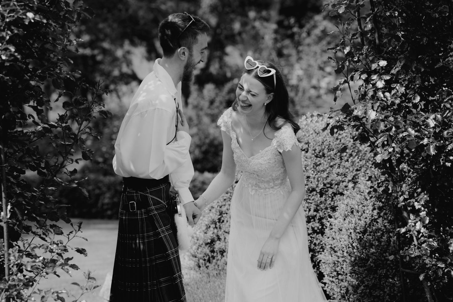 Laughing bride and kilt-wearing groom hold hands among green foliage in a black and white wedding photo.