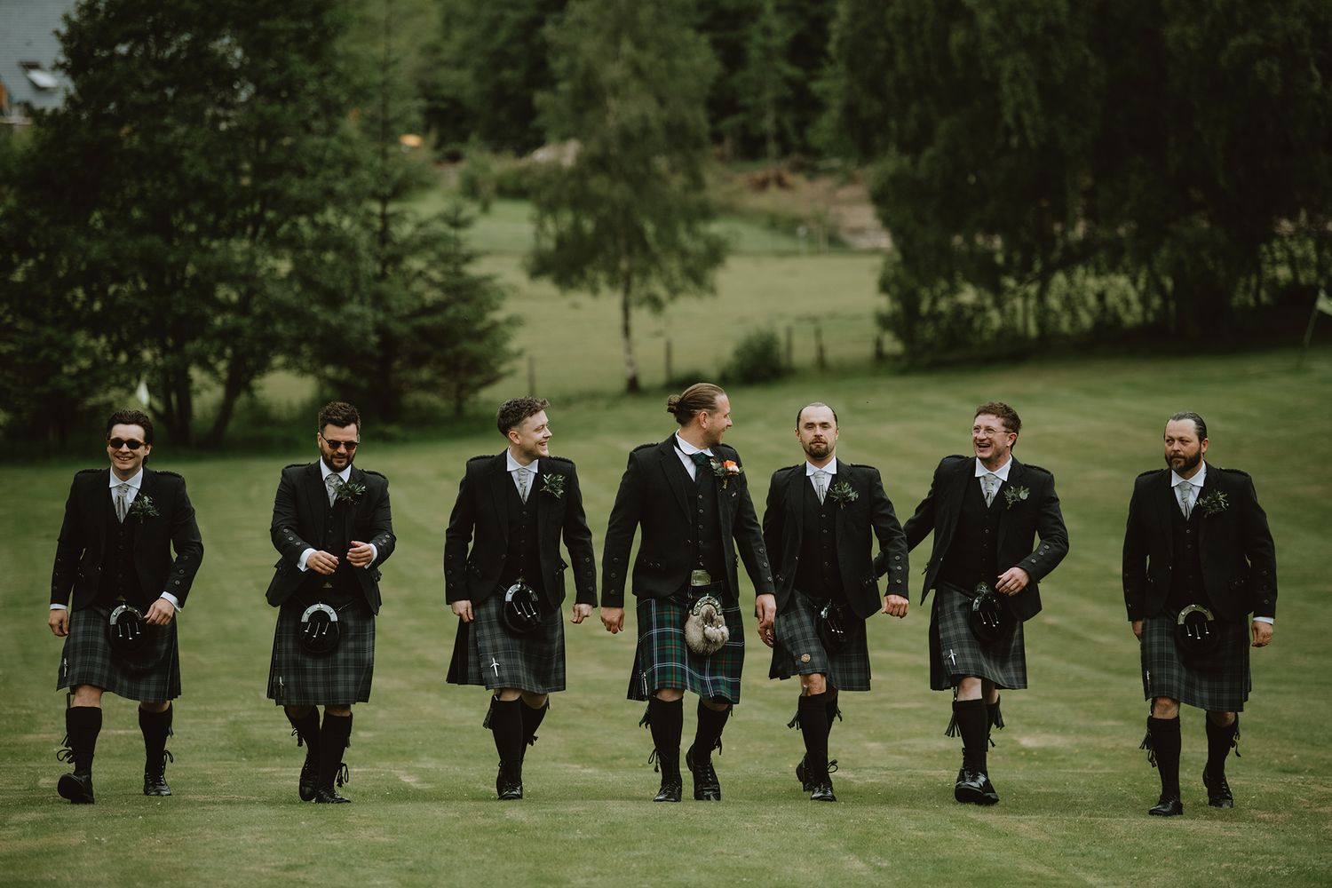 Seven groomsmen in kilts and suits walking across green field at Scottish wedding.
