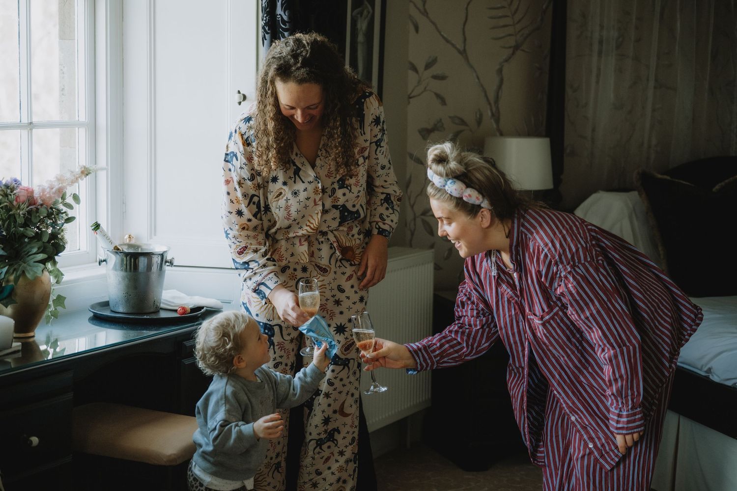 Mother in floral robe shares champagne toast with elderly woman and toddler in cozy wedding suite.