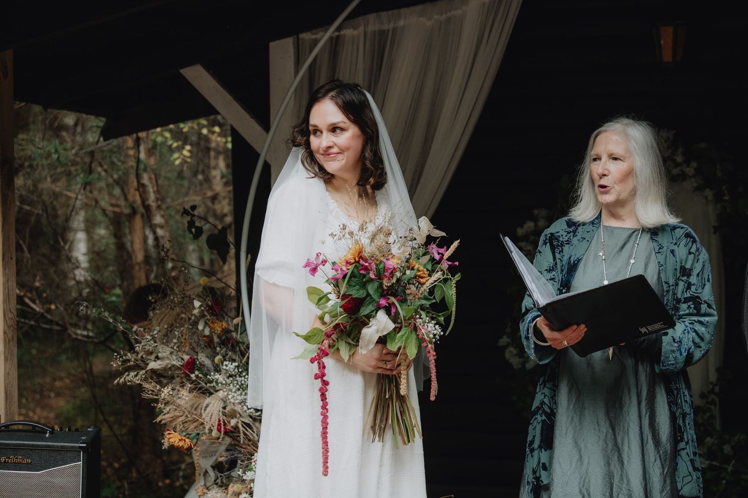 Smiling bride in white gown and veil holds colorful bouquet during outdoor ceremony.